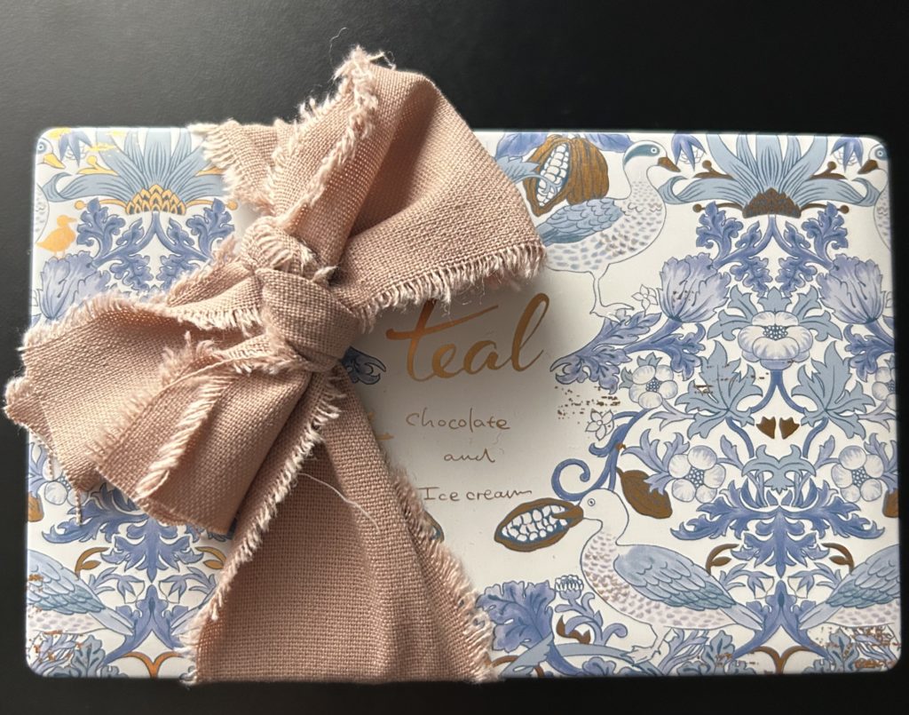 ウィリアム・モリスの「いちご泥棒」をモチーフにしたクッキー缶を、薄茶色のリボンで結んでいる。缶のデザインは、白い背景に青い線で描いたイラスト。苺はカカオに、小鳥は鴨に変わっている