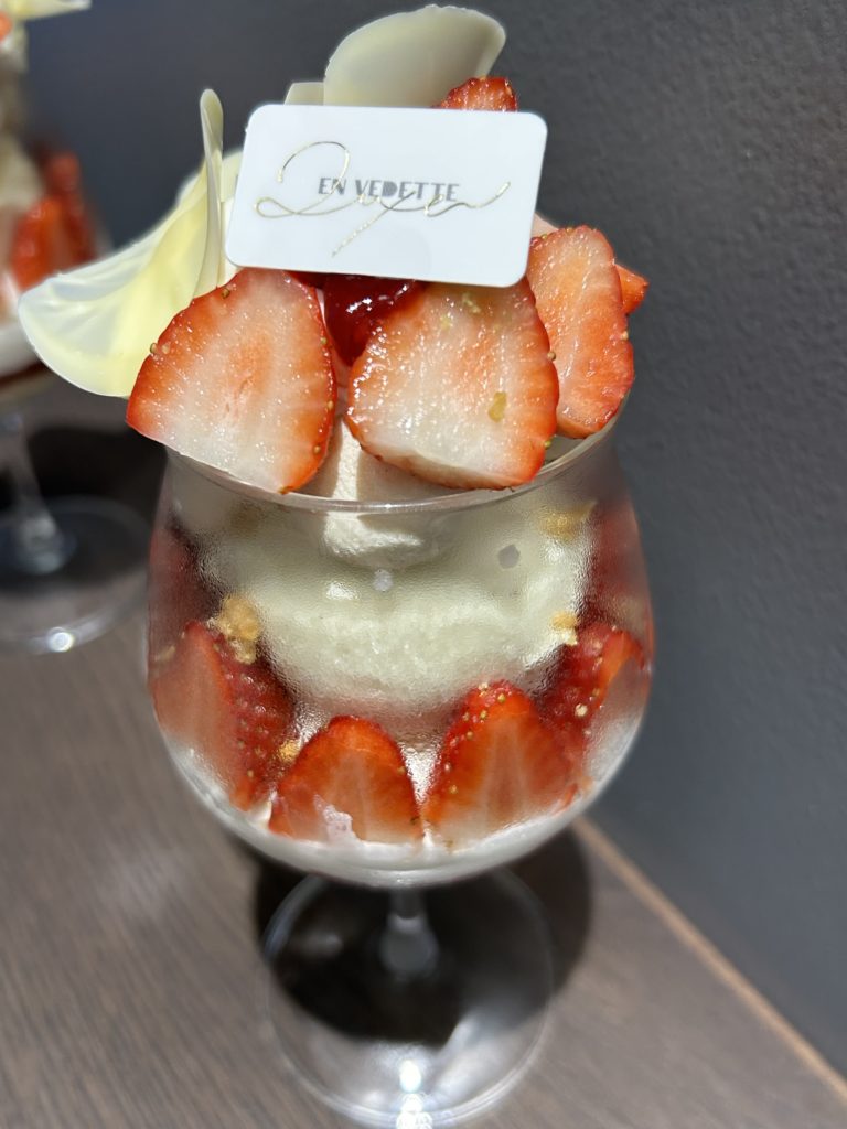 ショートケーキを模したパフェ。半分に切った苺を並べた上に、大きなバニラアイスと丸く絞った生クリームが覗き、その上にもう一度切った苺が並び、苺のソース(ジュレ?)や花びらのようなホワイトチョコを添えている。