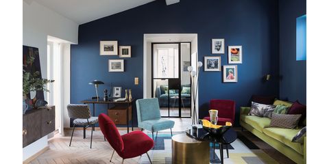 リビング奥側。奥の客室が見えるガラス戸の周りを青い壁が囲み、左右にそれぞれ写真か絵画が飾られている。手前に置かれたボルドーや水色の椅子が目を惹き、床や壁際のローソファの明度が高いのもあるからか、それほど暗い印象を与えない。