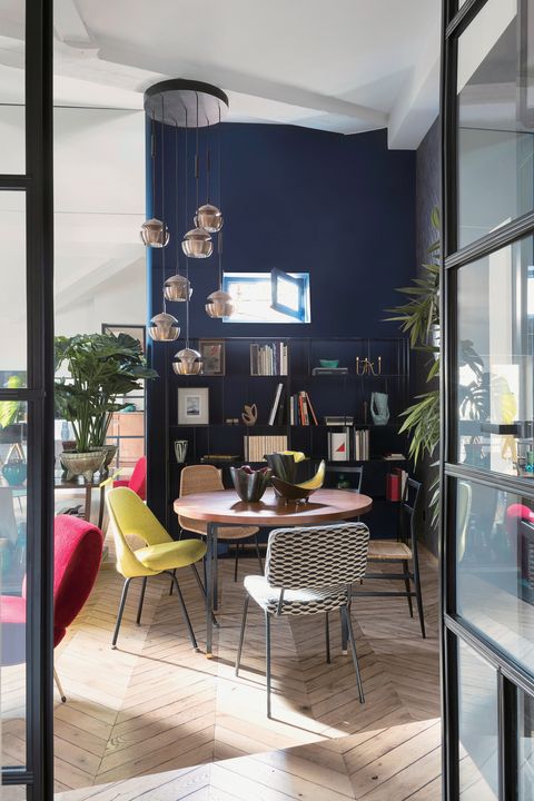 リビング（キッチンから見た図）。深い青色の壁沿いに置かれた黒色のラックには、本やオブジェ、絵などが飾っている。その手前にたくさんのペンダントライトがランダムに吊り下げられており、その下には丸テーブルを、足の細いカラフルな椅子が取り囲んでいる。
