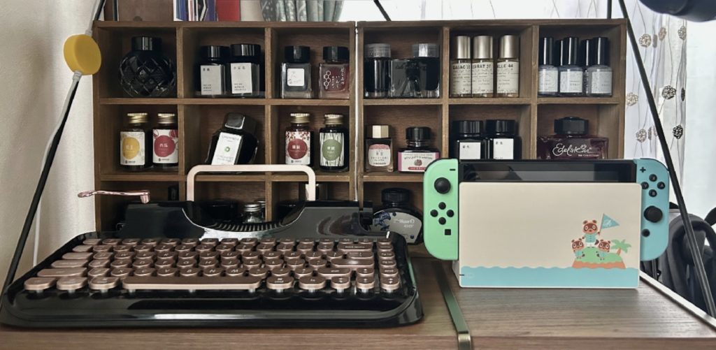 インク瓶を置いた木の箱の前に、黒とピンクゴールドのタイプライターとSwitch（とび森版）を置いた図。