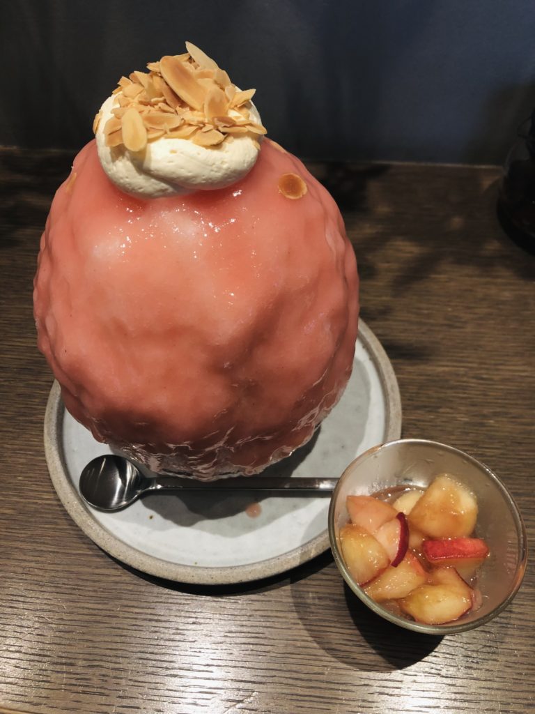 桃のかき氷。こんもり盛り上がった氷に濃いピンクの桃のソースがかかり、その上に白いクリーム、スライスアーモンドが乗っている。手前に小さな器で、味変用のカットされた桃が添えられている。