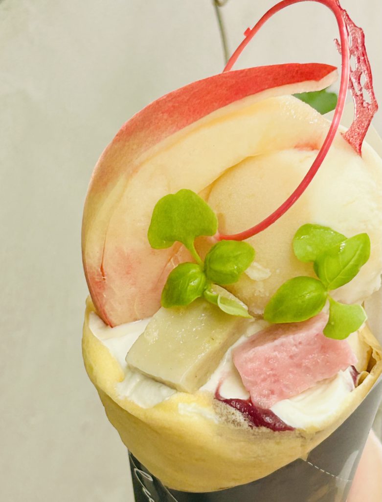 桃のクレープ。丸く包んだクレープ（白いクリームの土台）に、ピンクと薄緑の四角い麩、ハーブ、丸く大きな桃のソルベ、くし切りの桃。桃とソルベの間に輪っか状の飴が飾られている。