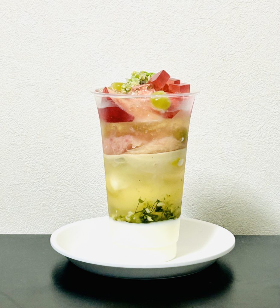 パルフェ ア アンポルテペシュ（横から見た図）
下からブラマンジェの白、フェンネル、レモンジュレ等の透けた黄色、白餡やディスクショコラの白、切り分けられた桃、下のジュレより黄味が濃いジュレの上、ローズヒップ寒天の濃いピンクやずんだ餡、うぐいす豆の黄緑で彩られている。