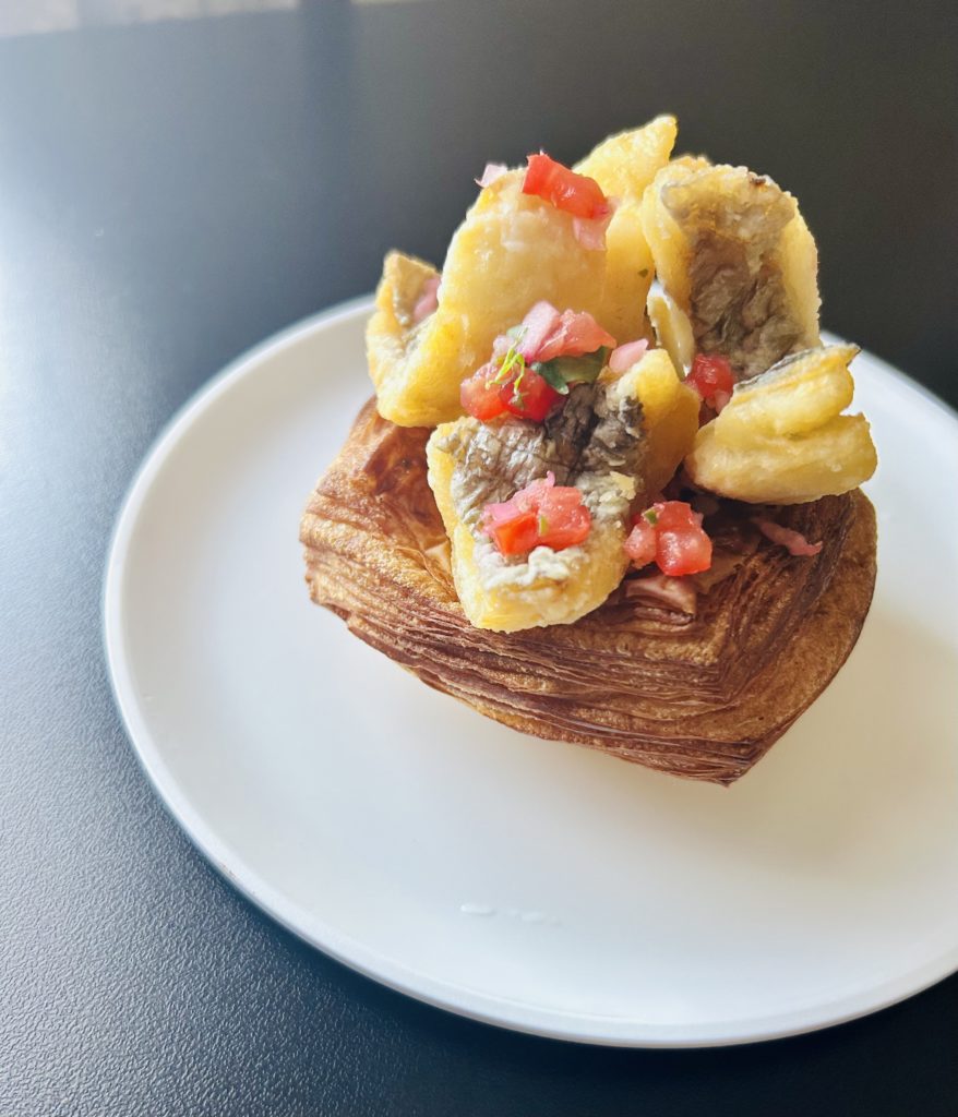 太刀魚のフリット サルサソースデニッシュ。四角いデニッシュ生地の上にU字型に曲がった太刀魚のフリットが4切れ乗せられ、その上にハーブ、みじん切りしたトマトと紫玉ねぎが飾られている。