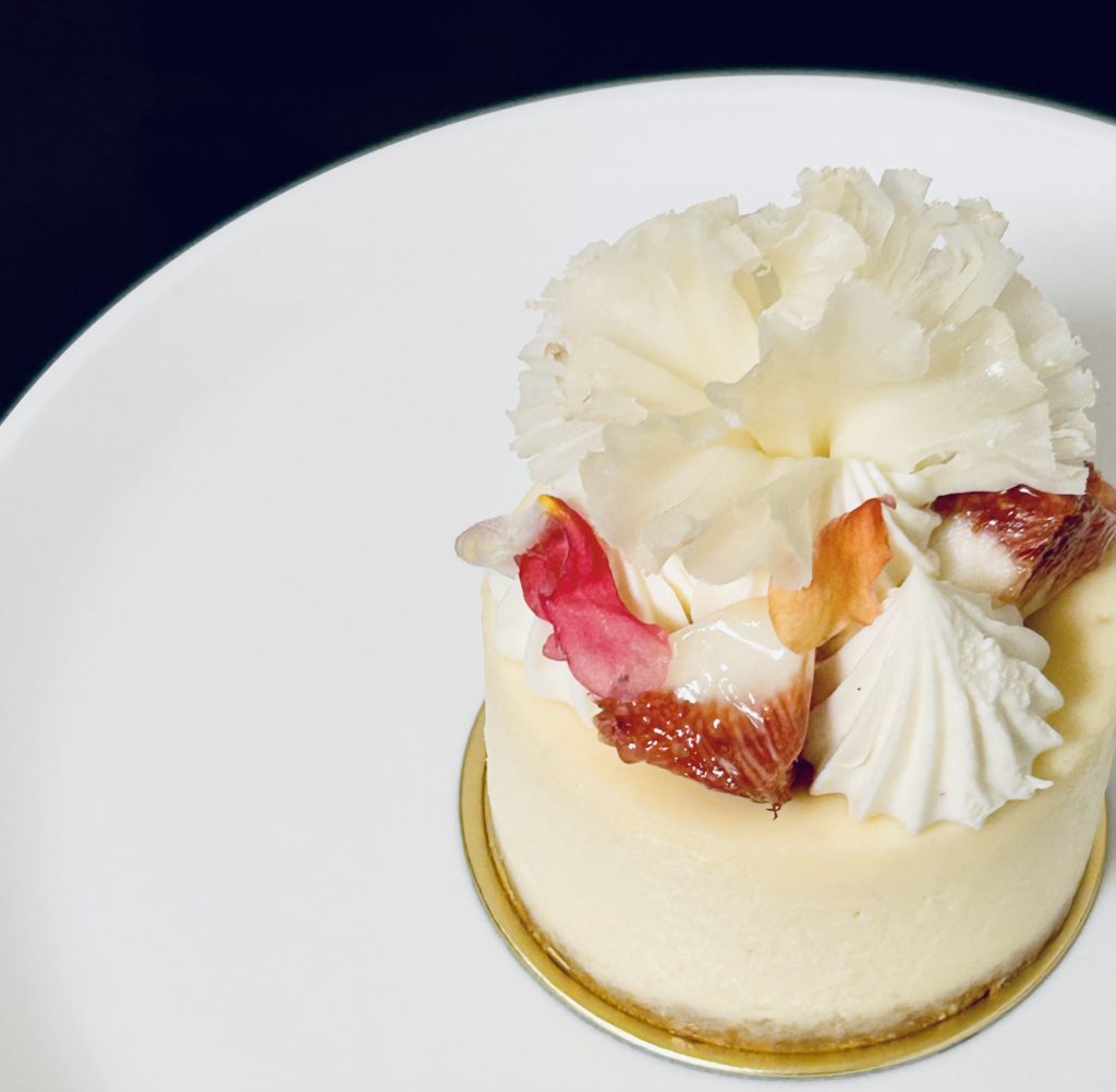 ゴルゴンゾーラのチーズケーキ。円柱型の白いケーキの上に、カットしたいちじく、生クリーム、赤やオレンジの花びら、カーネーションみたいに削ったチーズが乗っている。