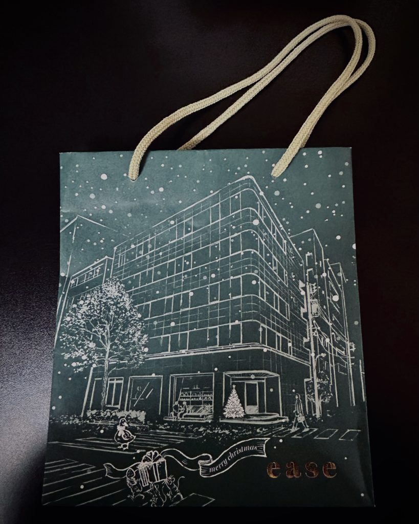 easeの紙袋。深緑の紙に、店舗の入ったビル、ツリー、外で菓子を食べる二人、横断歩道を渡る鴨やプレゼントを運ぶネズミ達。